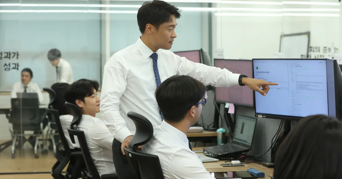 사무실에서 한 사람이 모니터 화면을 손가락으로 가리키며 앉아 있는 동료 둘에게 설명하고 있다